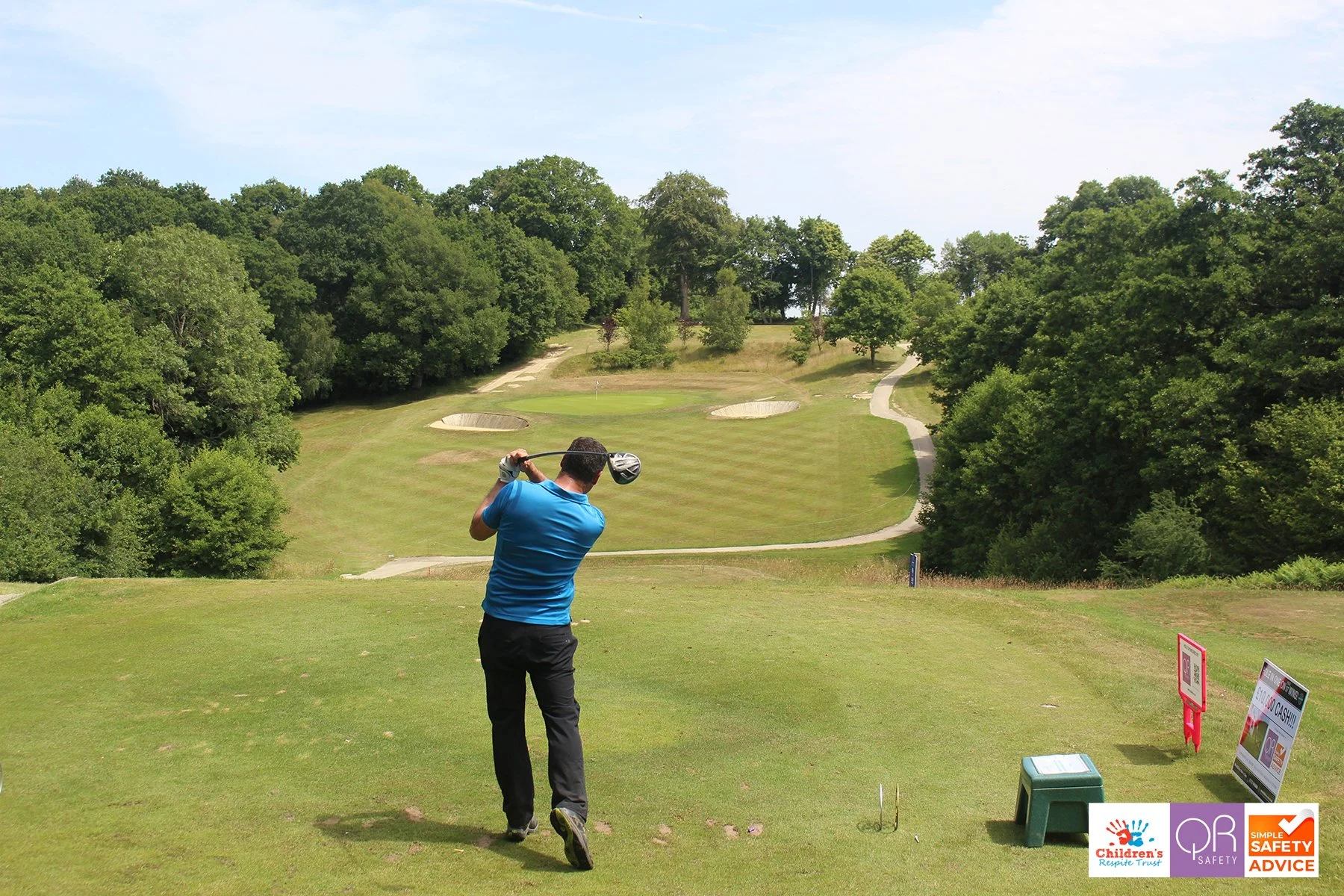 A man in a blue shirt and black pants swinging a golf club on a golf course surrounded by trees, with sand traps and a flag in the distance.