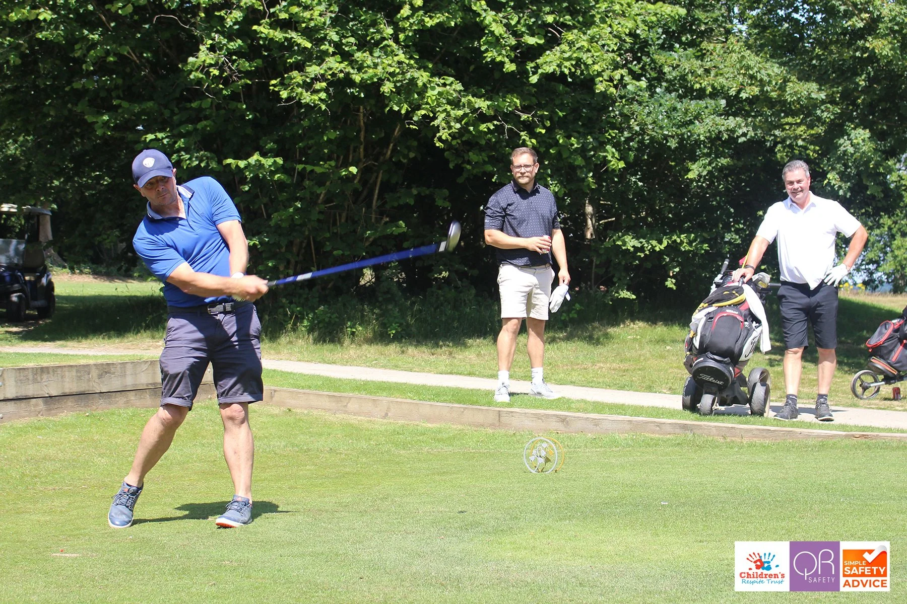 A man in a blue polo shirt and shorts swings a golf club on a golf course while two men in casual clothing watch nearby, with golf bags in the background.
