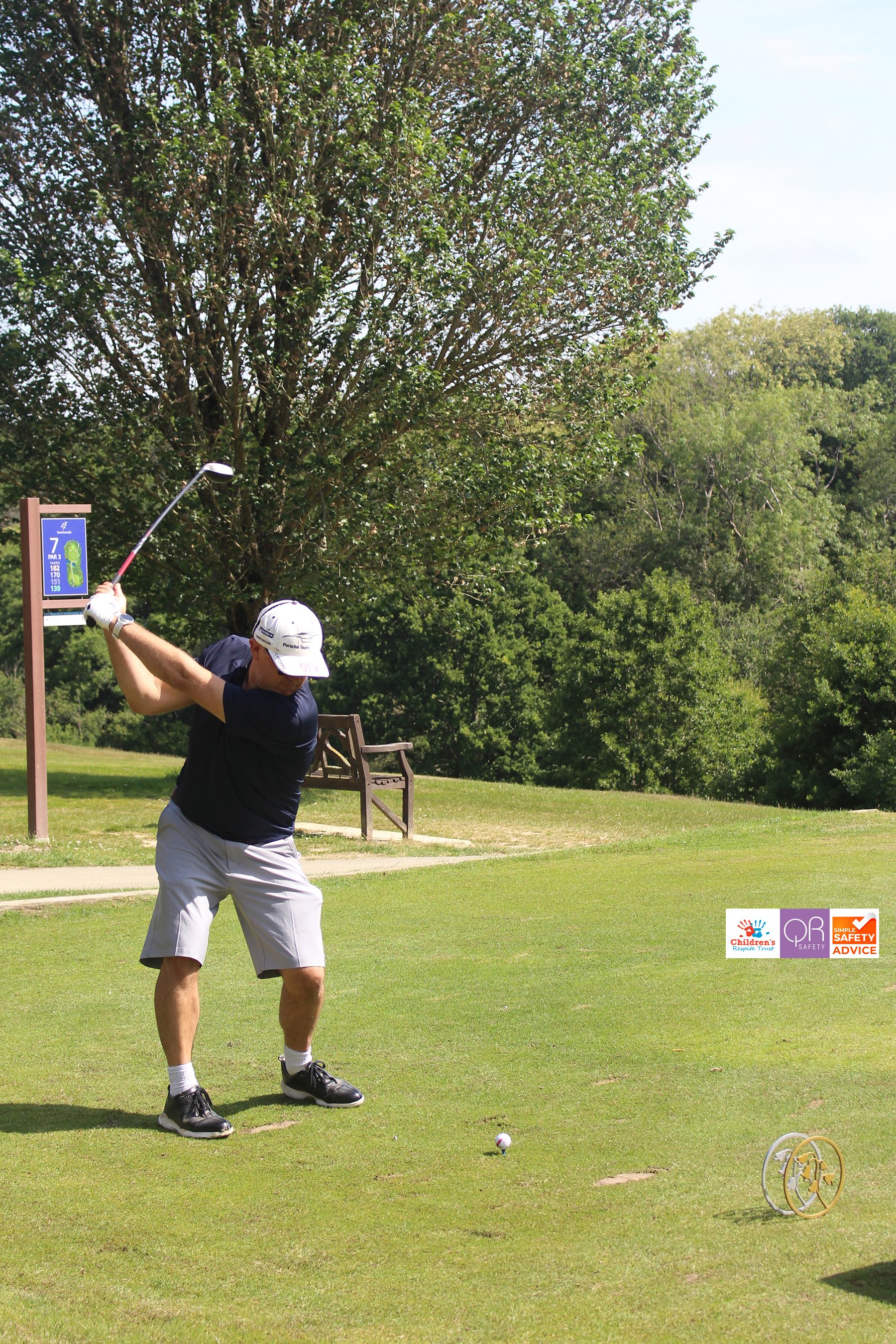 Man playing golf on a green golf course, swinging a club. There is a large tree in the background, a bench nearby, and signs along the course.