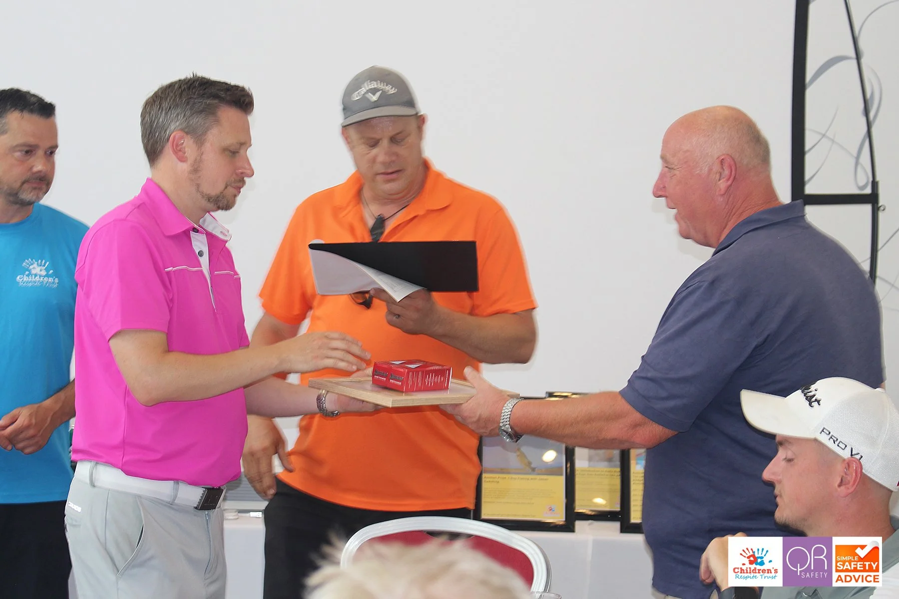 A man in a pink shirt is receiving an award on a wooden plaque with a red box on top from a man in a dark blue shirt. Three other men are observing, one in an orange shirt holding a clipboard, another in a blue shirt and a white cap, and one in a lig