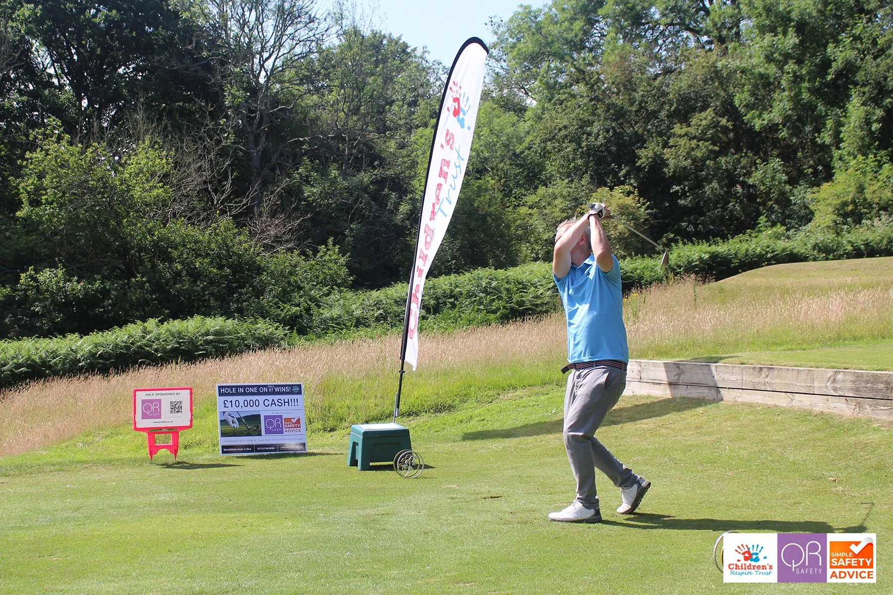 A man playing golf on a green course, swinging a golf club with a wooded background and promotional signs nearby.