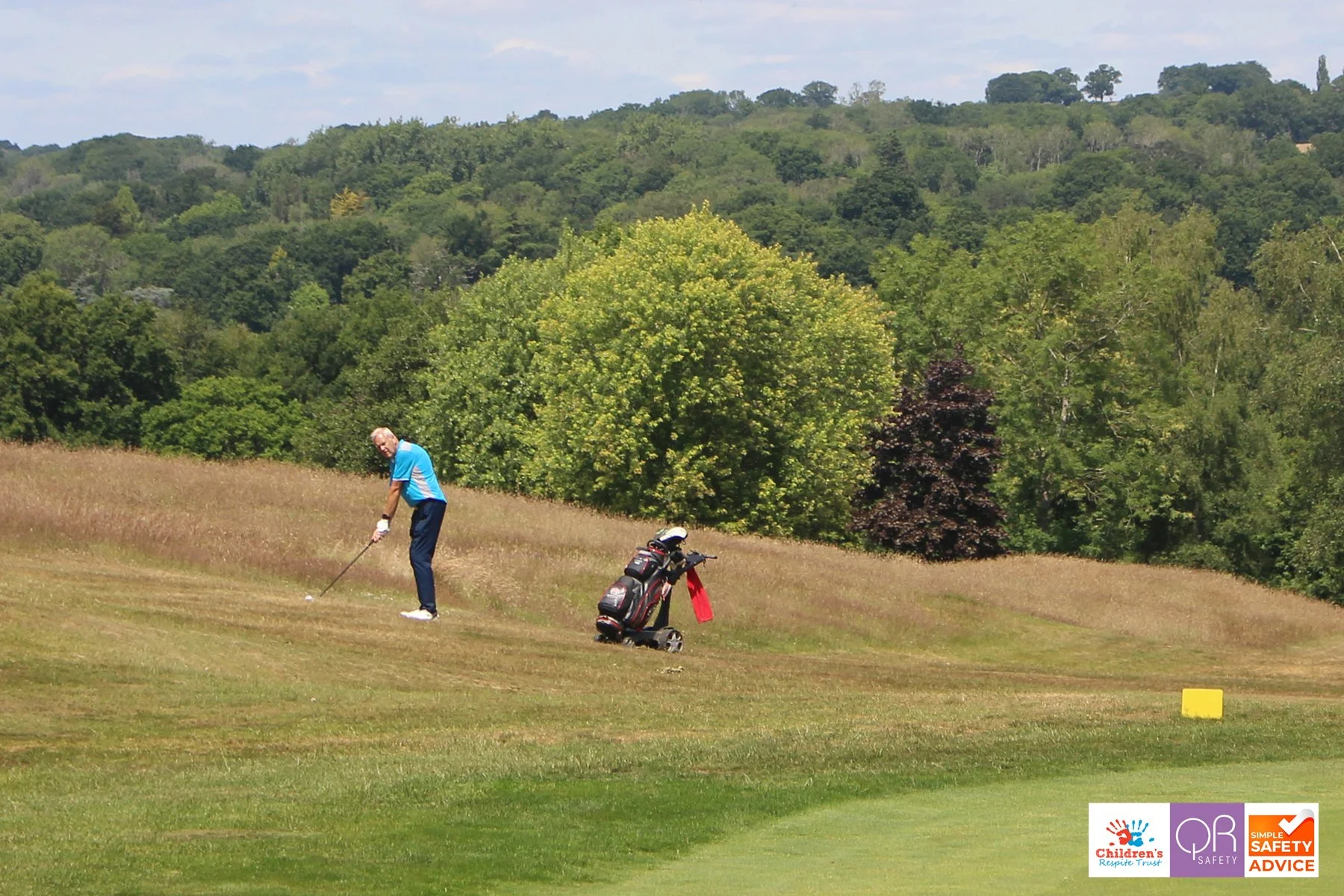 A man playing golf on a lush golf course with multiple trees and hills in the background. The man is dressed in a blue shirt and dark pants, swinging a golf club. There is a golf bag with clubs nearby and safety and children’s trust logos in the bott