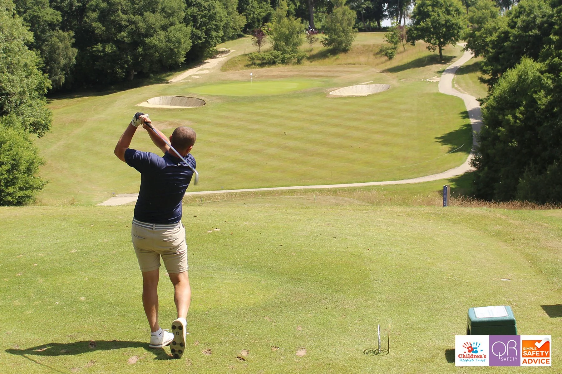 A man is swinging a golf club on a golf course with green grass, trees, and sand bunkers.