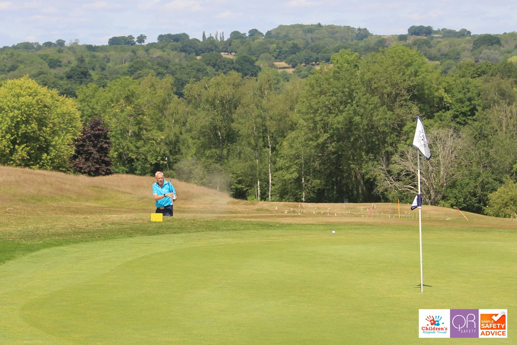 A person on a golf course preparing to hit a golf ball towards the hole, with a flag on a green, surrounded by trees and hills in the background.