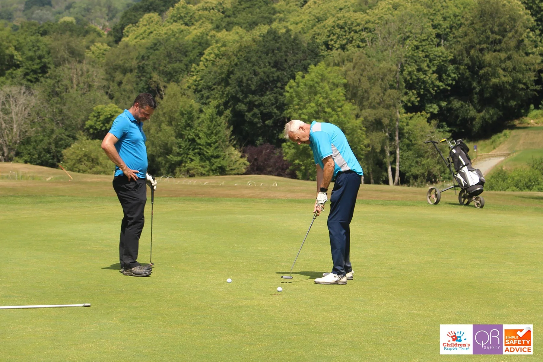 Two men on a golf course practicing putting, one with a golf club and the other observing, with a golf cart and lush green trees in the background.