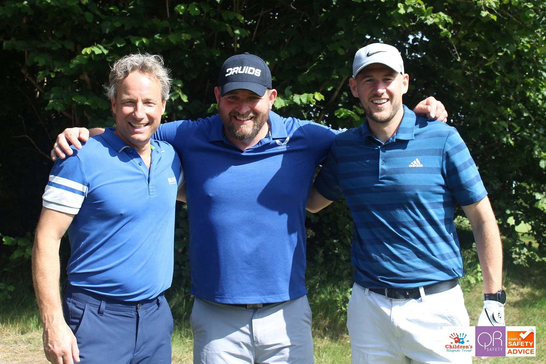 Three men standing outdoors in front of green trees, smiling with arms around each other, dressed in blue golf shirts and hats, and one wearing a golf glove.