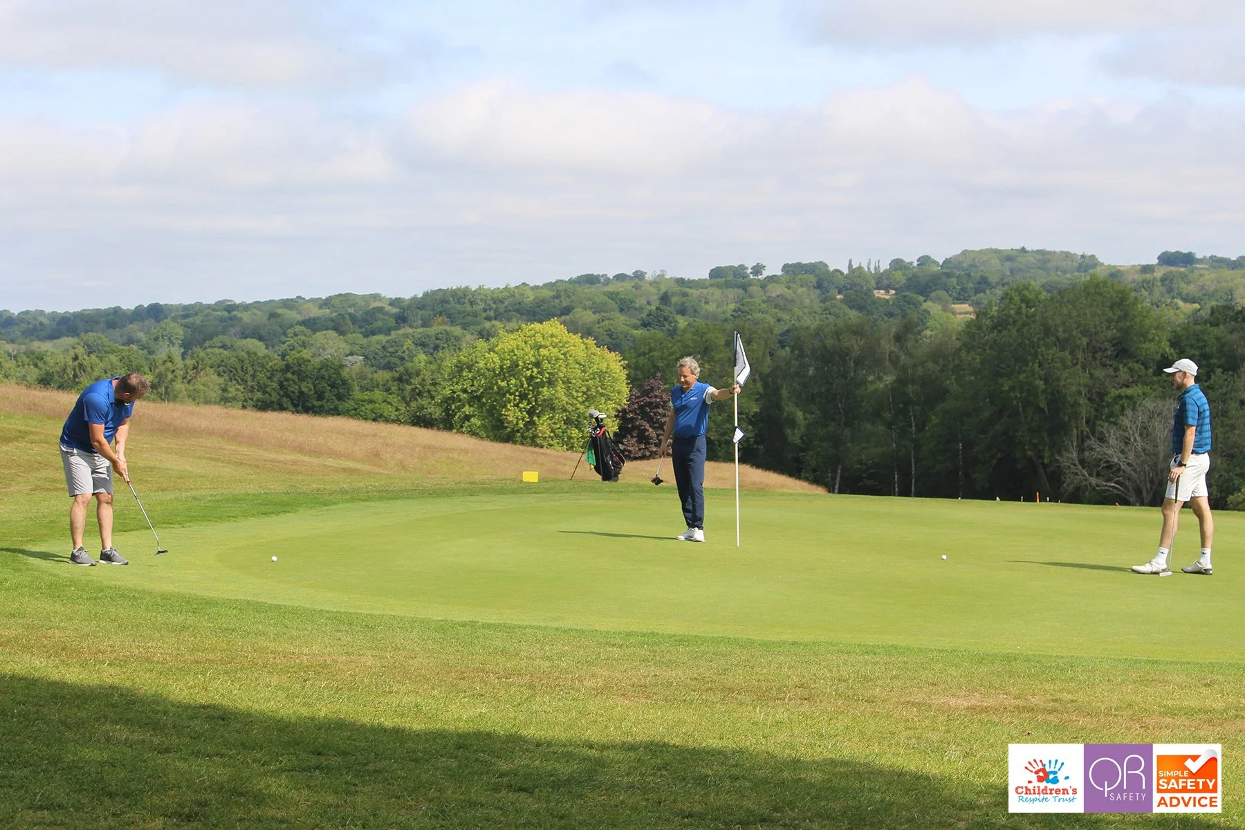 Three men playing golf on a lush green course with trees and hills in the background on a partly cloudy day.