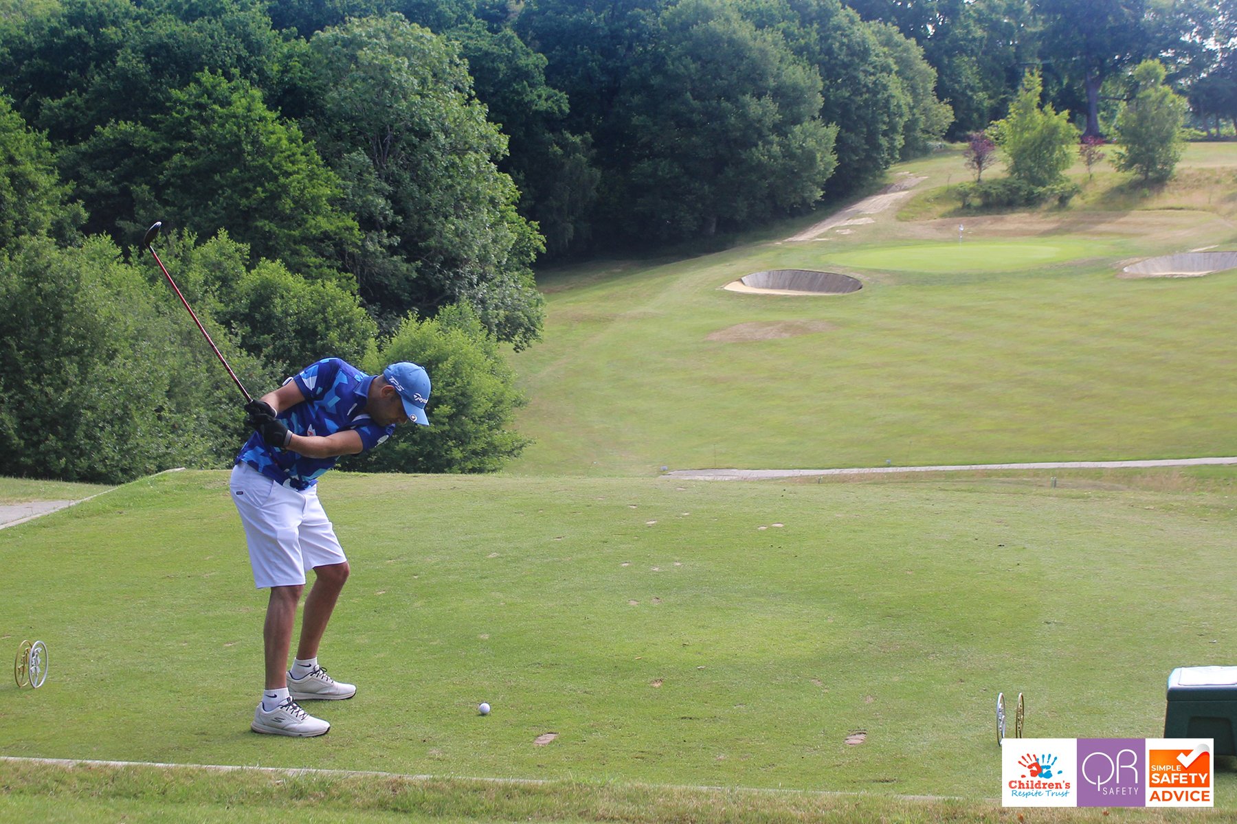 Young male golfer in blue shirt, white shorts, and cap preparing to hit the golf ball on a grassy tee box with trees and sand bunkers in the background.
