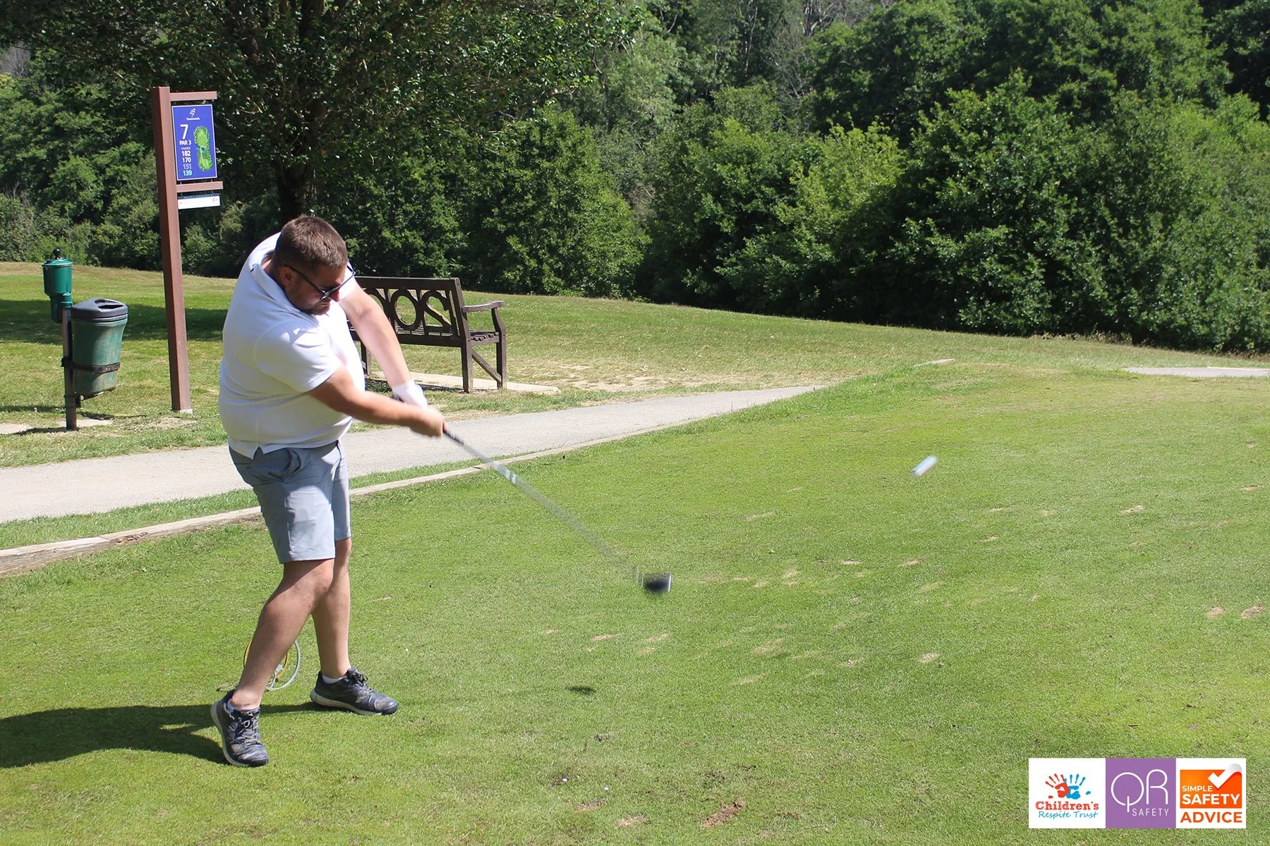 A man wearing glasses, a white t-shirt, and gray shorts swings a golf club on a golf course. There are trees, a bench, a sign indicating hole 7, and trash cans in the background.