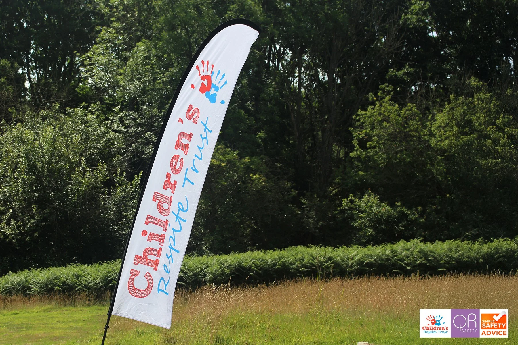 A vertical banner reads 'Children's Respite Trust' in red, blue, and black lettering, placed outdoors on a grassy field with tall grass, bushes, and trees in the background.