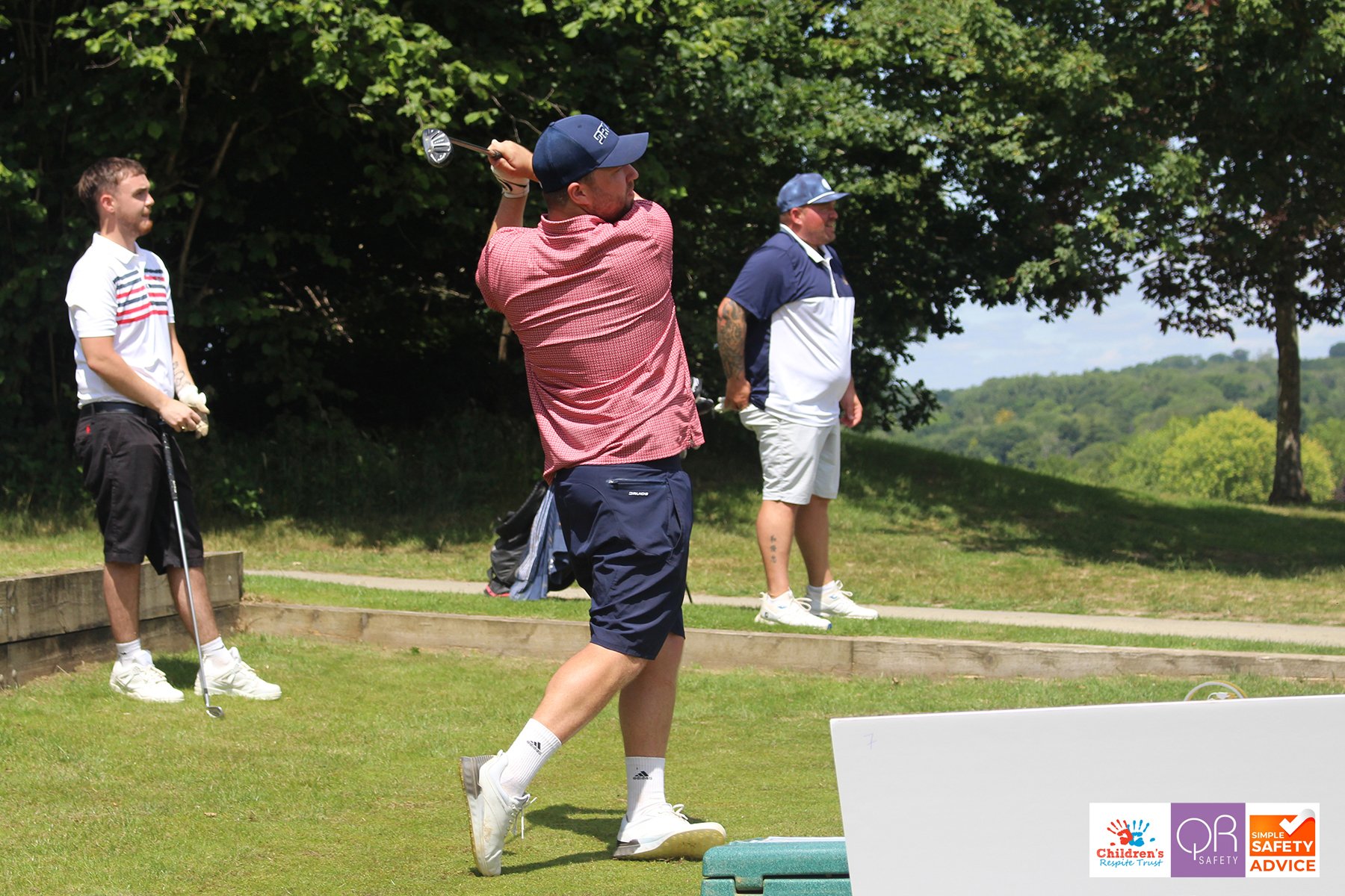 Three men on a golf course, one swinging a golf club while the others watch, with trees and a blue sky in the background.