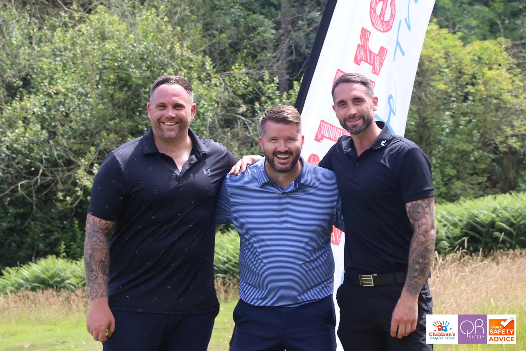 Three smiling men standing outdoors, hugging each other, with a banner in the background. They are dressed in casual sportswear, and the background is a natural, green setting.