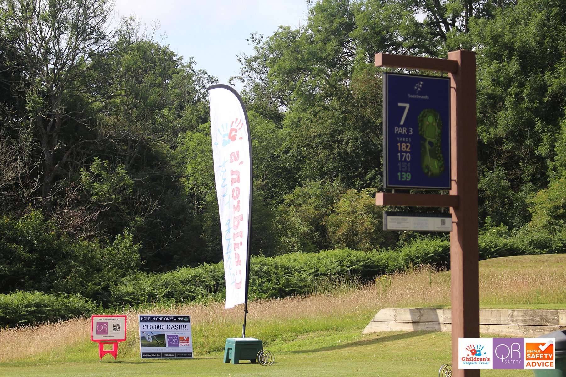 A golf course in the daytime with multiple signs, banners, and advertisements, including a tall blue sign showing hole number 7 and par 3, distances, and a children's charity banner near trees and grass.