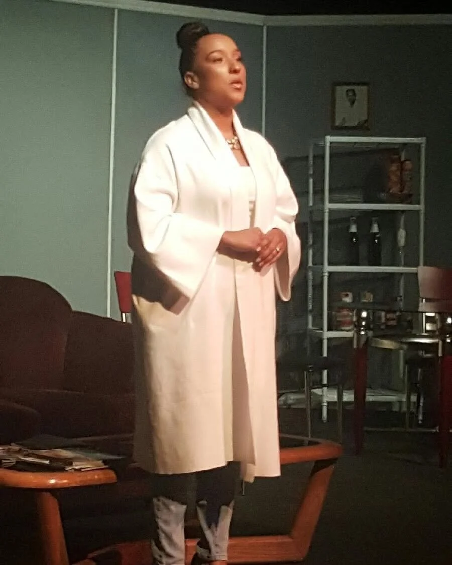 A woman dressed in a white coat and black pants, standing in a room with shelves and chairs, possibly during a performance or presentation.