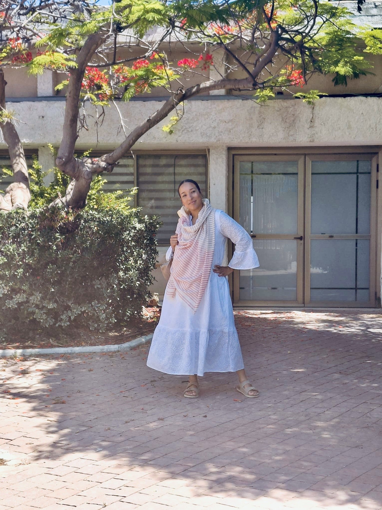 A woman standing outdoors in front of a building with large windows and a sliding glass door. She is wearing a white dress, a pink and white striped shawl, and sandals. There is a tree with red flowers and green leaves shading her from the sunlight.