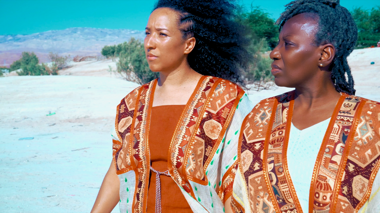 Two women standing outdoors on a sandy landscape, wearing traditional patterned garments, with a desert-like background and a mountainous horizon, looking to the side.
