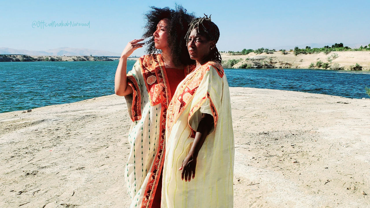 Two women wearing colorful traditional clothing standing near a body of water on a sandy shore with hills in the background.