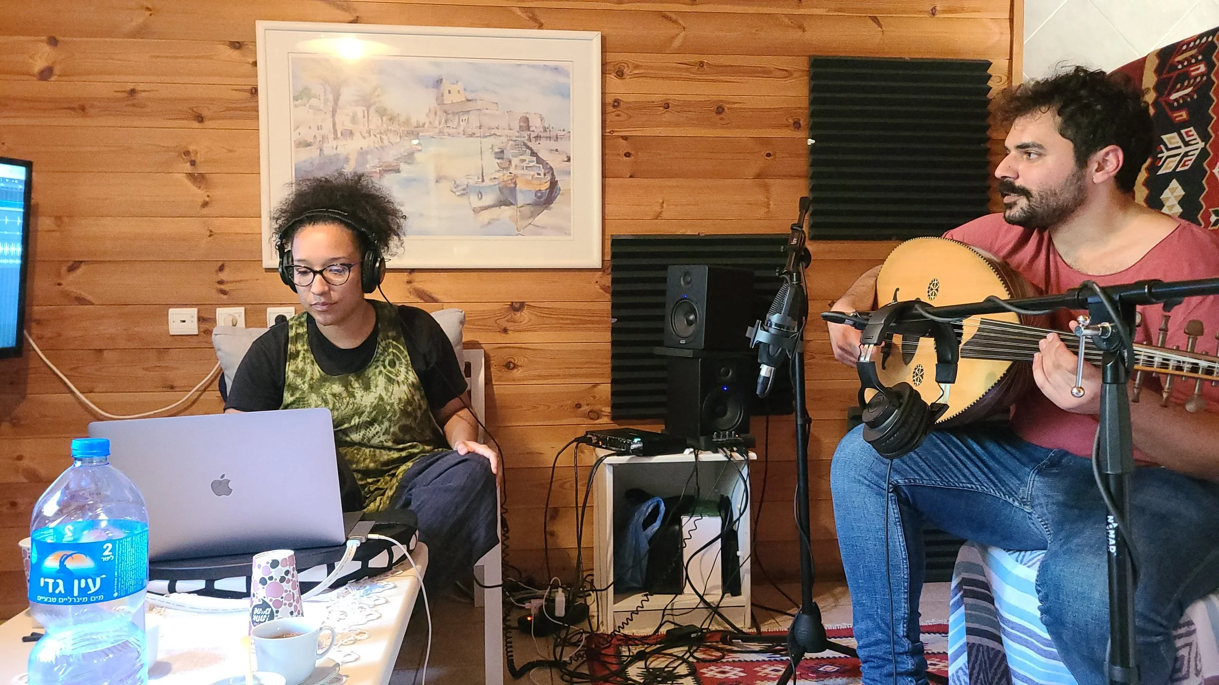 Two people in a music studio: a woman with curly hair, glasses, and headphones working on a laptop, and a man with dark hair and beard playing an acoustic guitar, with wall-mounted soundproofing and artwork of boats in the background.
