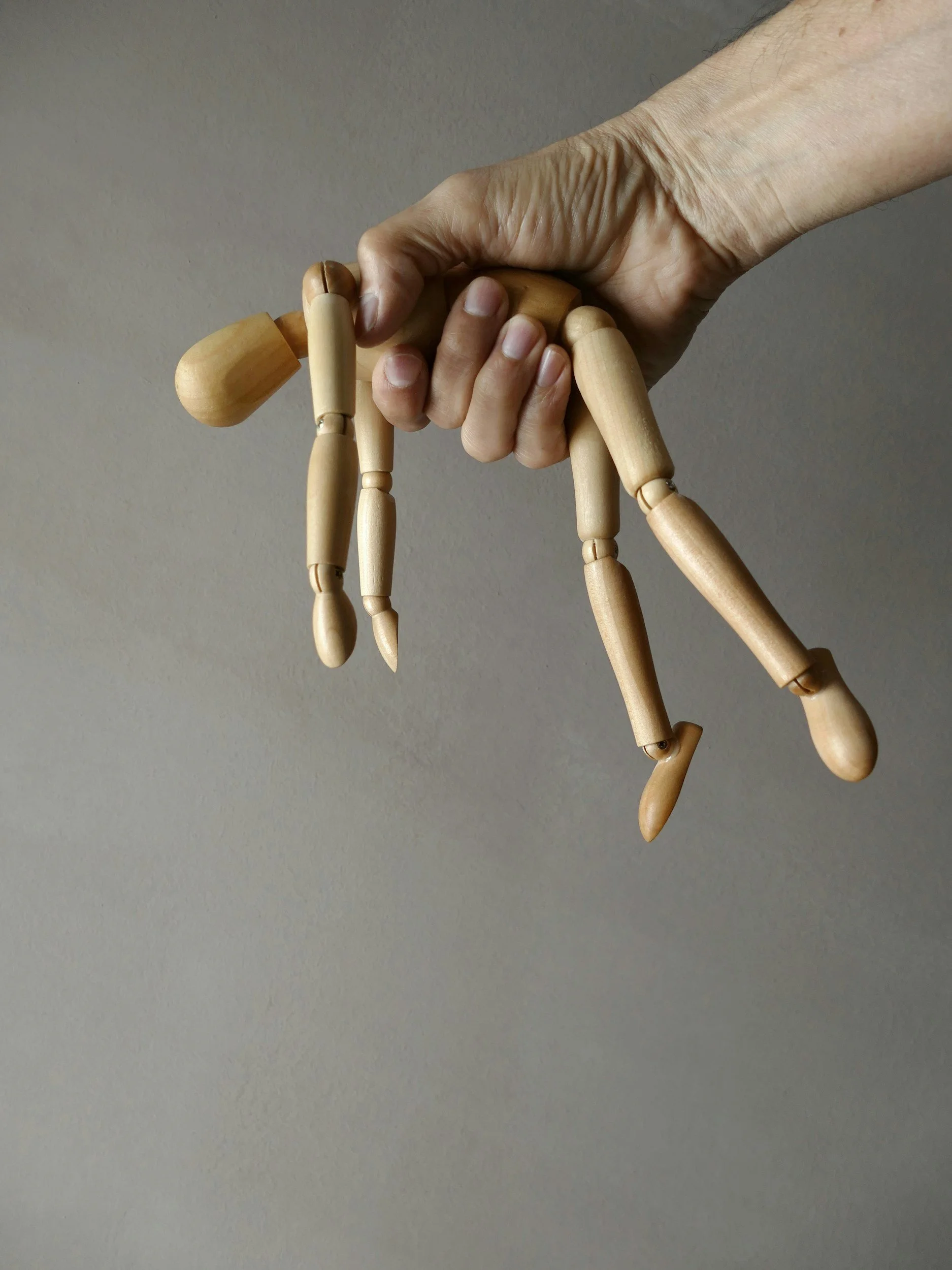 A person's hand gripping a wooden artist's mannequin's hand against a plain grey wall.