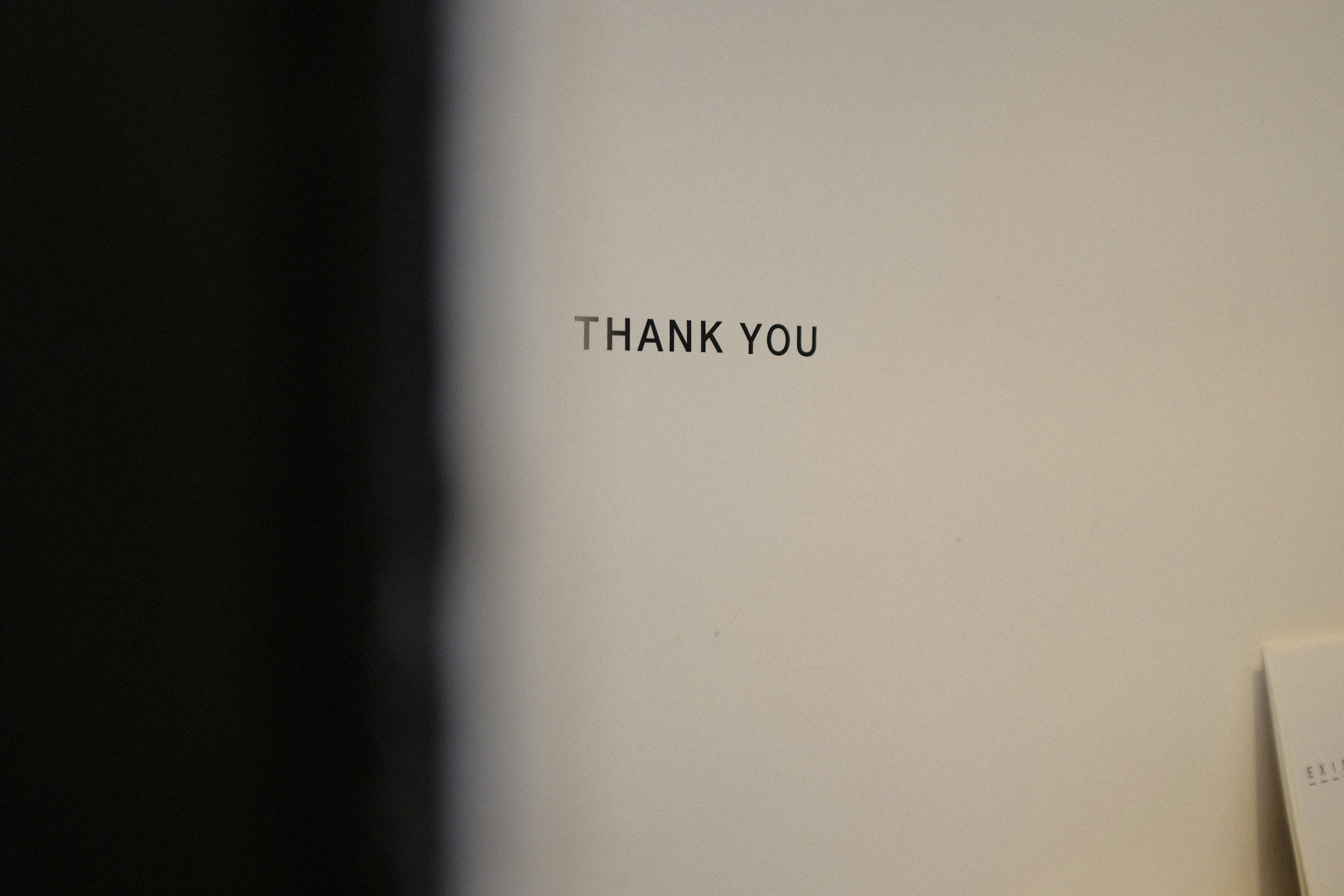 A close-up of a wall with the words 'Thank You' printed on it. The wall is mainly white with a dark shadow on the left side of the image.