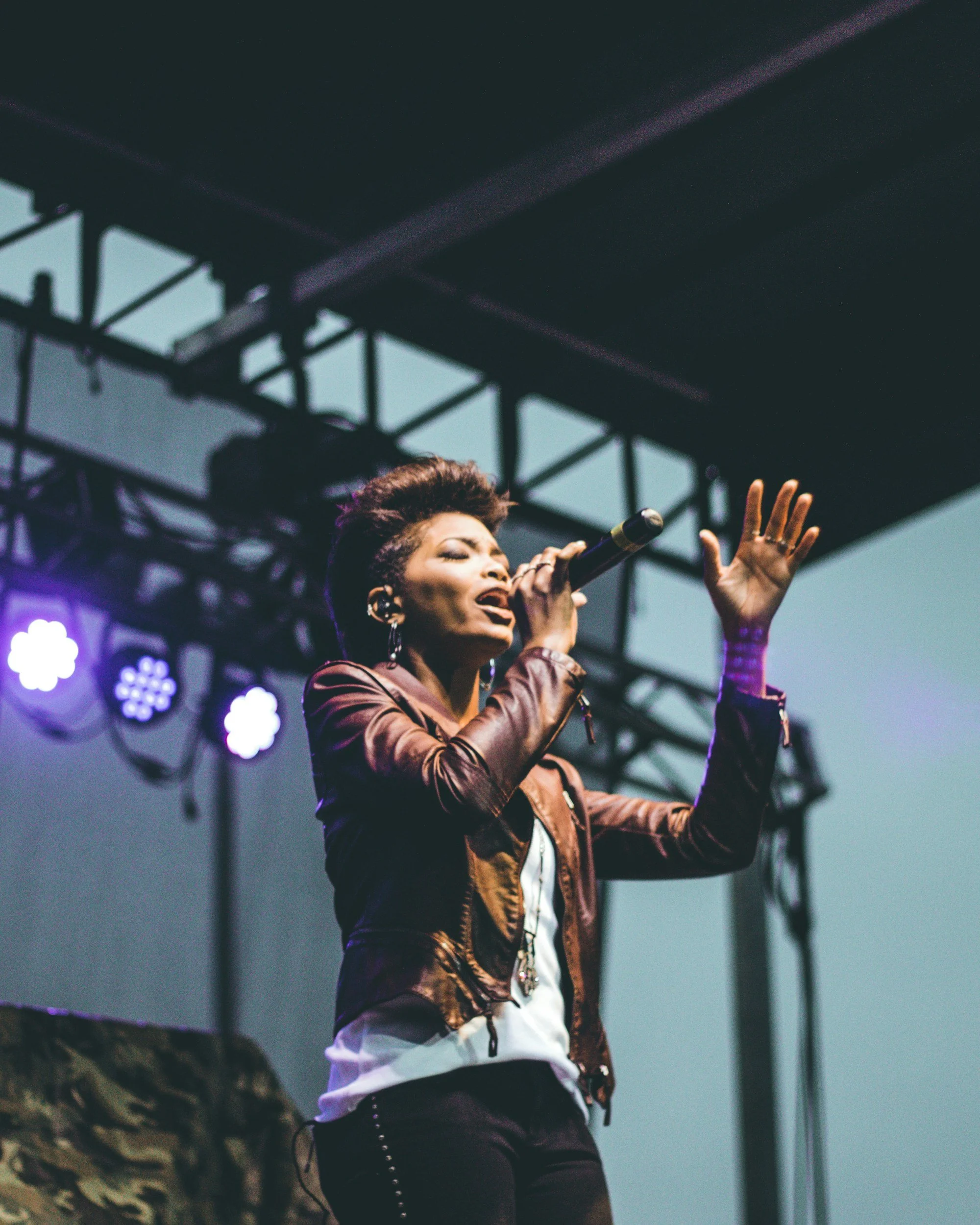 A female singer performs on stage with a microphone, wearing a brown leather jacket and black pants, gesturing with one hand, with stage lights and a metal framework overhead.