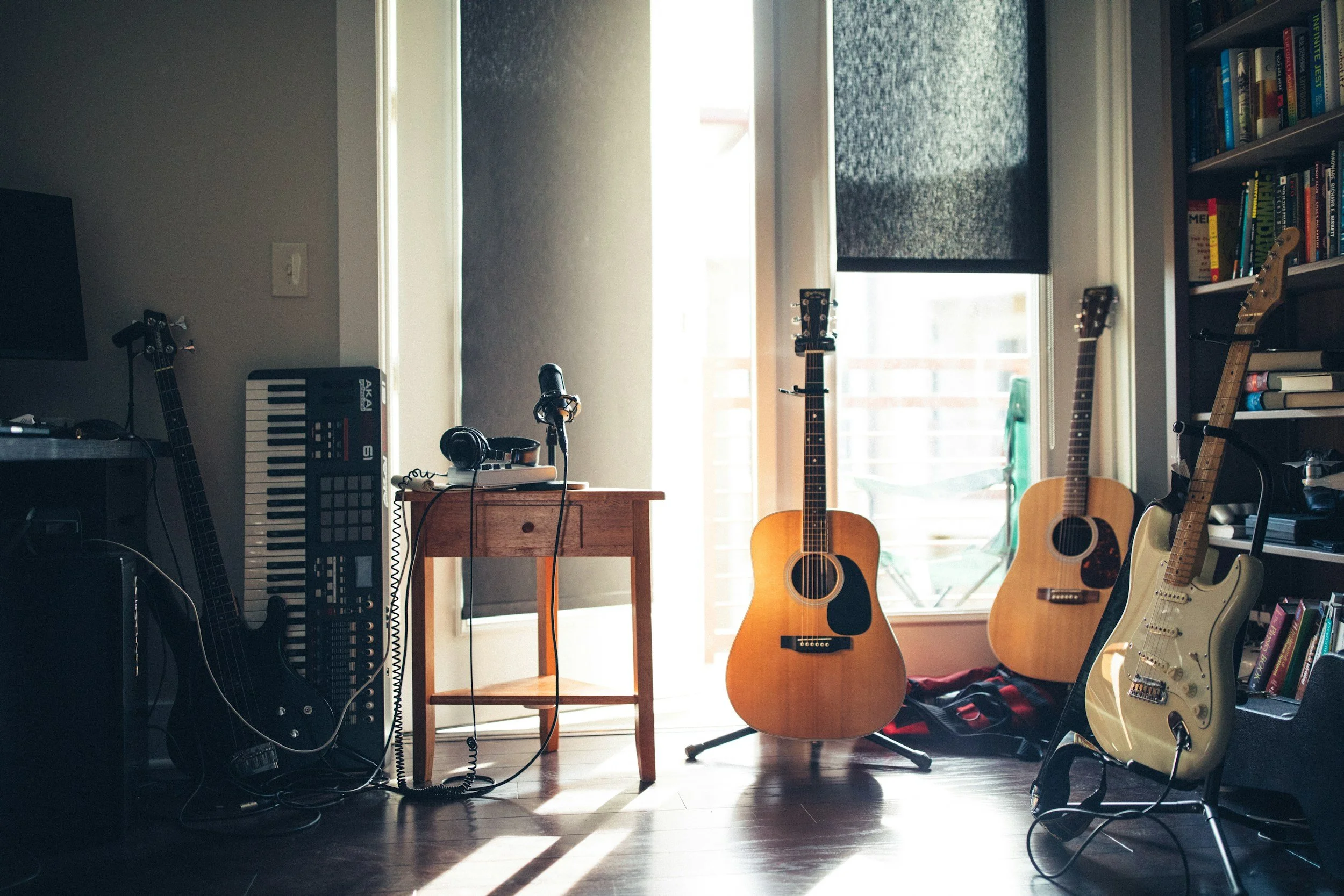 A music studio with guitars, a keyboard, a microphone, headphones, and a bookshelf near sliding glass doors with black curtains.