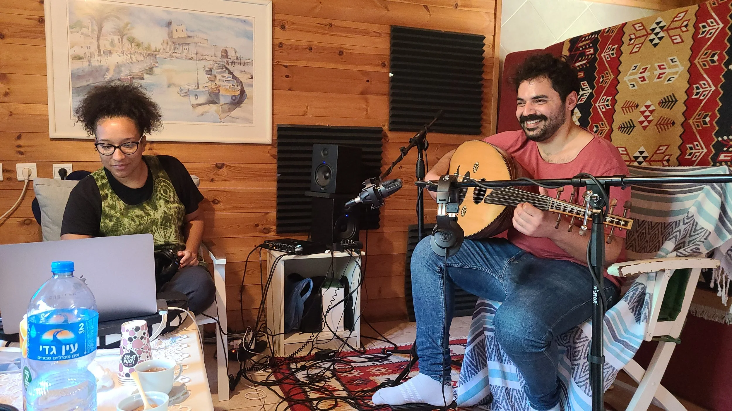 Two people are in a cozy room with wooden walls. One woman with glasses and curly hair looks down, possibly at a laptop. A man with a beard, smiling, is sitting on a chair playing an acoustic guitar. The room has a framed painting of boats on water, 