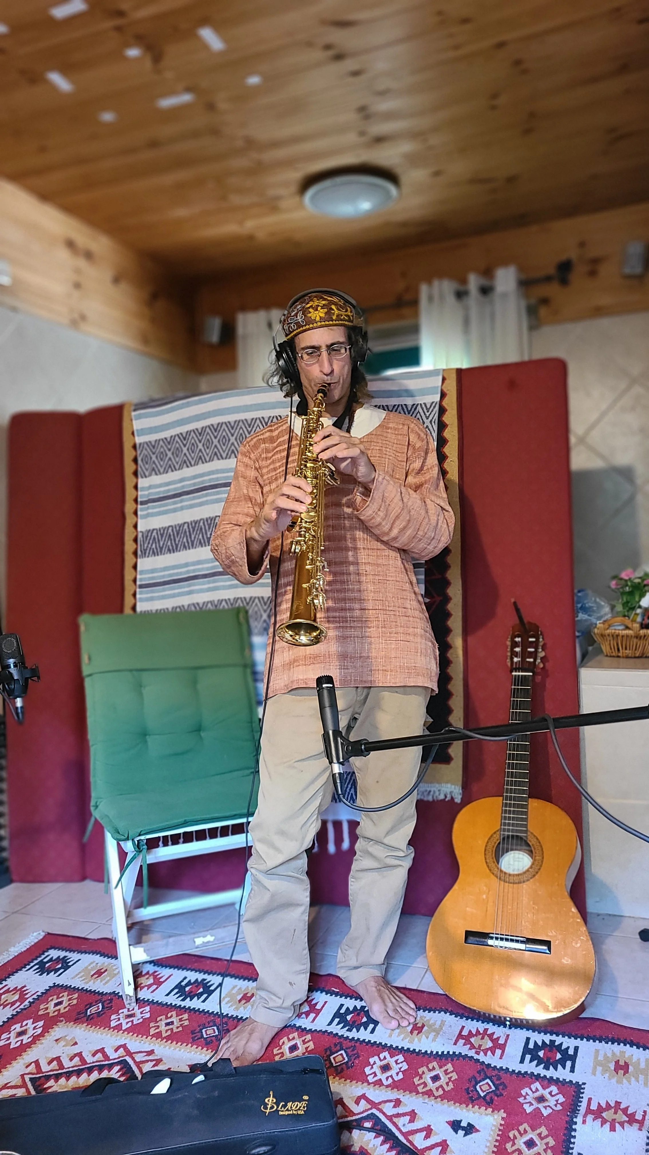 A man with long dark hair, glasses, and a decorative headband is playing a saxophone in a cozy room. He is wearing a traditional long-sleeved orange shirt and light-colored pants. Behind him, a guitar is leaning against the wall, and there are variou