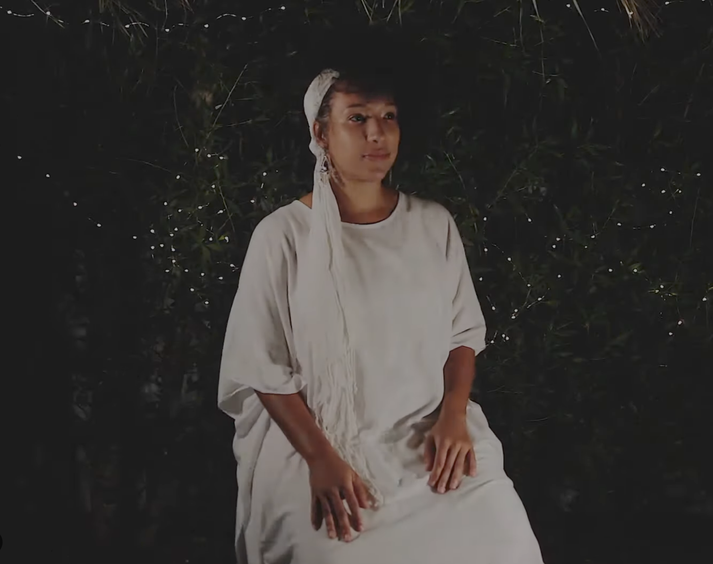 A person sitting outdoors at night, wearing a white long-sleeved shirt and a hat, with hands resting on their lap, surrounded by dark foliage and small lights in the background.