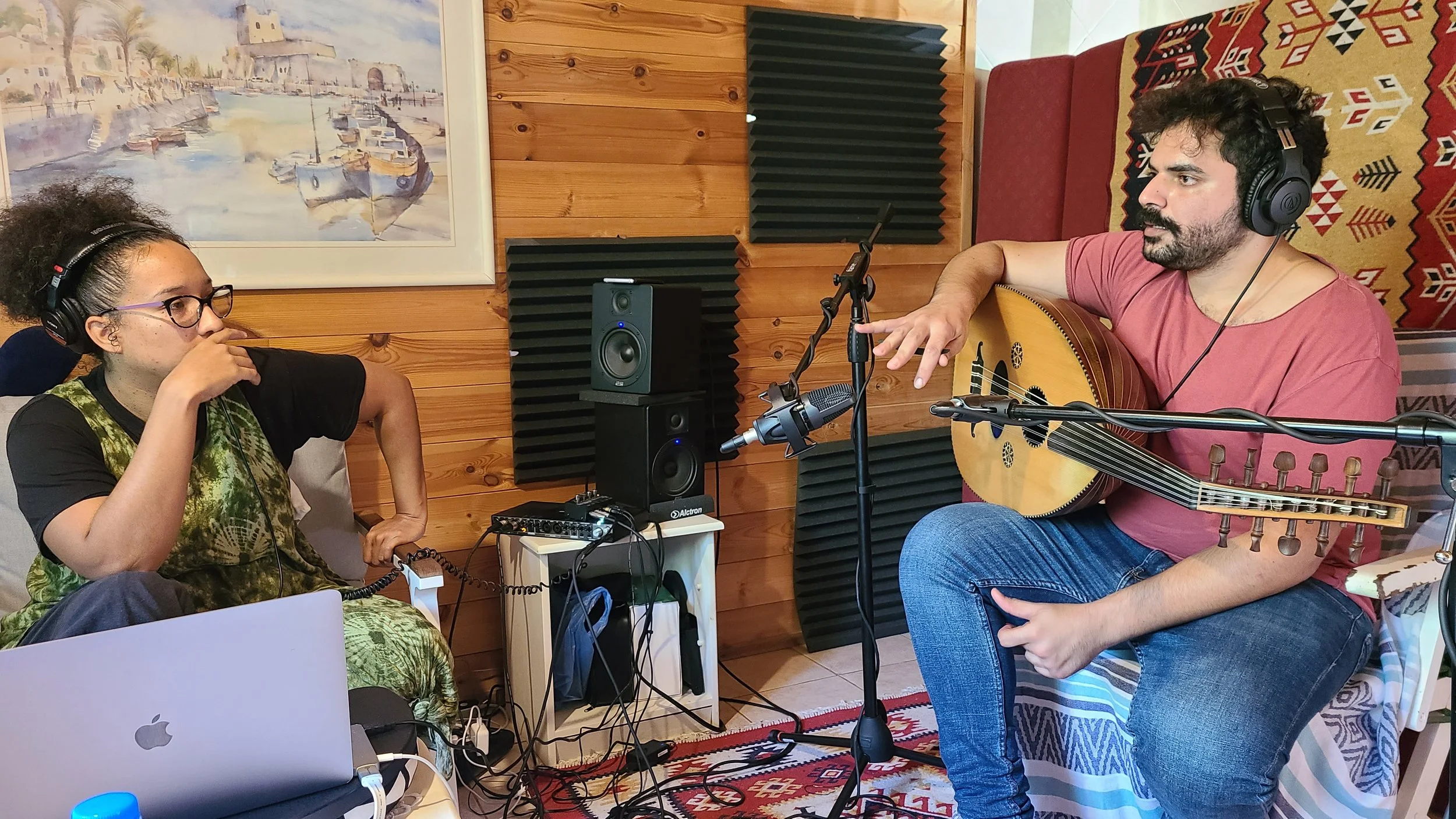Two people in a recording studio, one playing guitar and singing into a microphone, the other wearing headphones and listening, with recording equipment and acoustic foam on the wooden wall behind them.