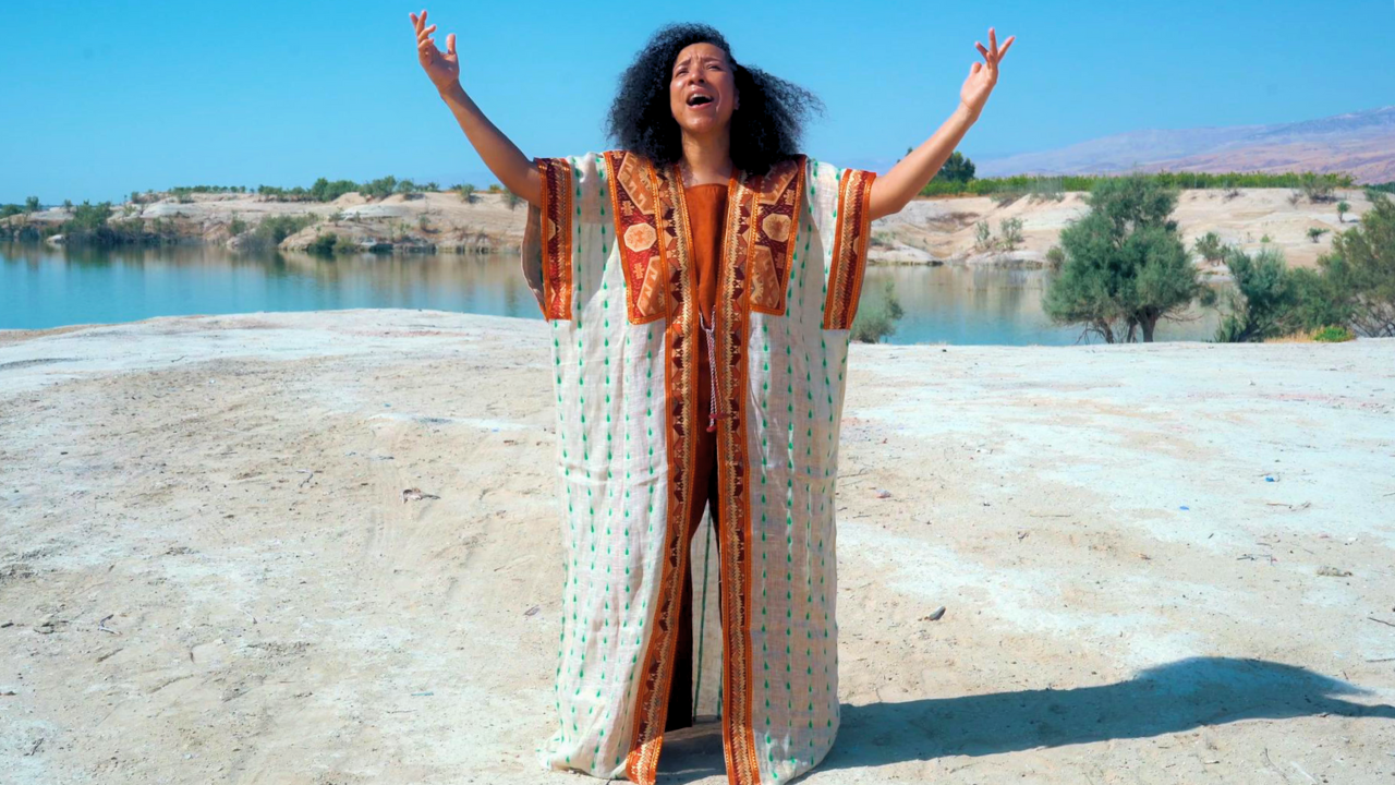 A woman with curly black hair wearing a colorful bohemian dress stands on a sandy beach with arms raised. Behind her is a river, green trees, and distant hills under a clear blue sky.