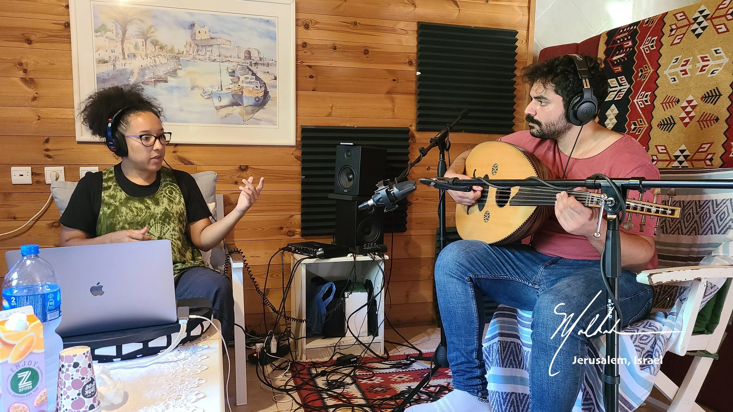 A woman wearing glasses and headphones sitting on a couch, speaking into a microphone, and a man playing an acoustic guitar, both wearing headphones, in a cozy room with wooden walls and framed artwork.