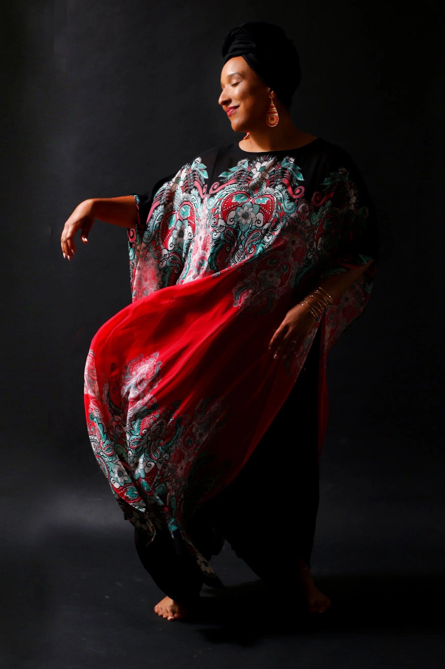 A woman dancing on a dark background, wearing a flowing, colorful patterned dress, a black headwrap, and jewelry, with her eyes closed and a smile.