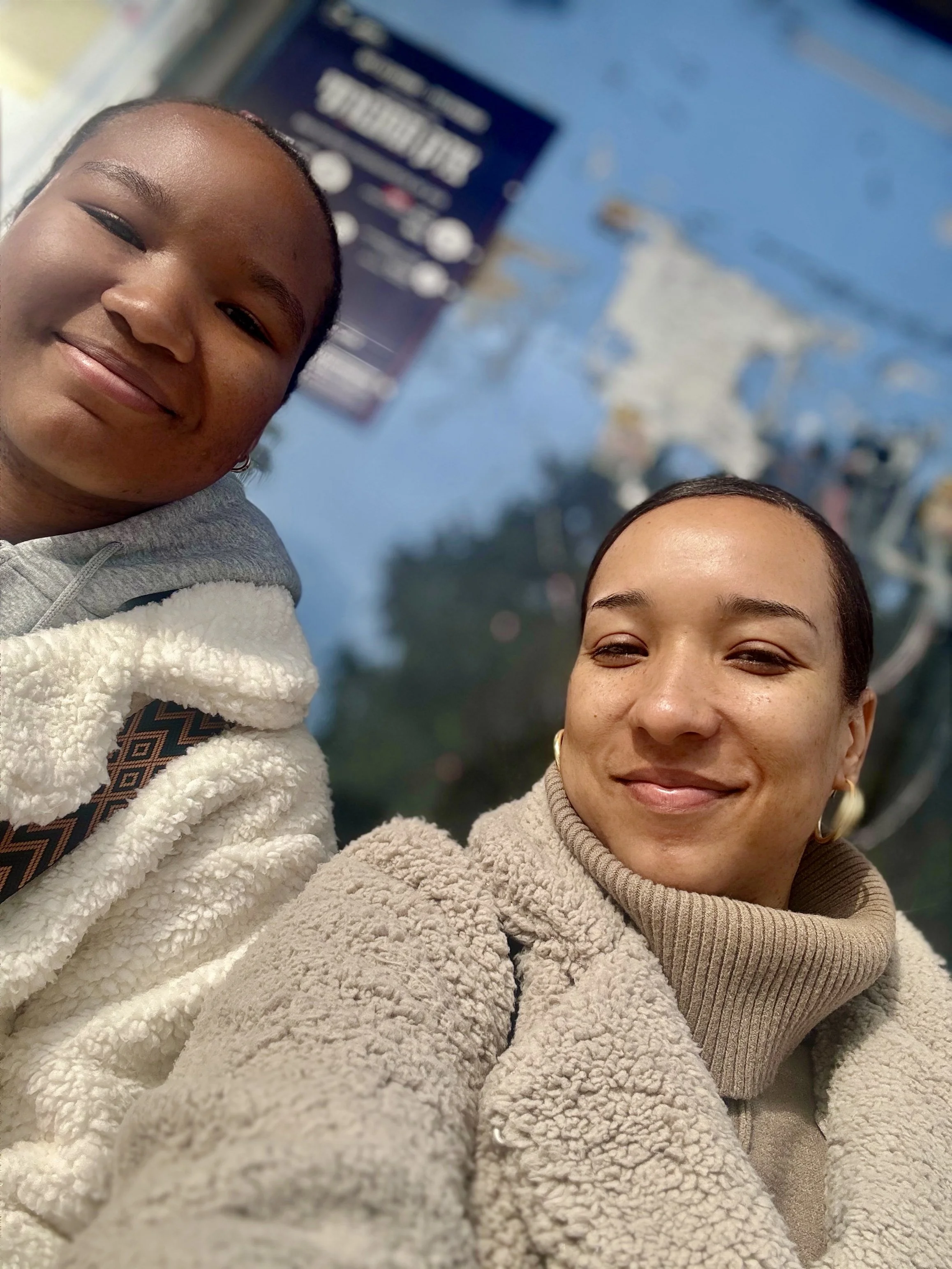 Two women taking a selfie outdoors, smiling, with a blue sky and amusement park rides in the background.
