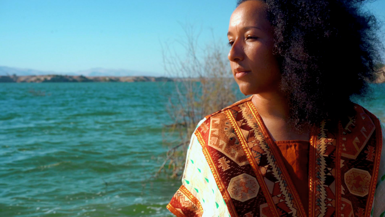 A woman with natural curly hair stands near a body of water, looking to the right, wearing a colorful patterned garment.