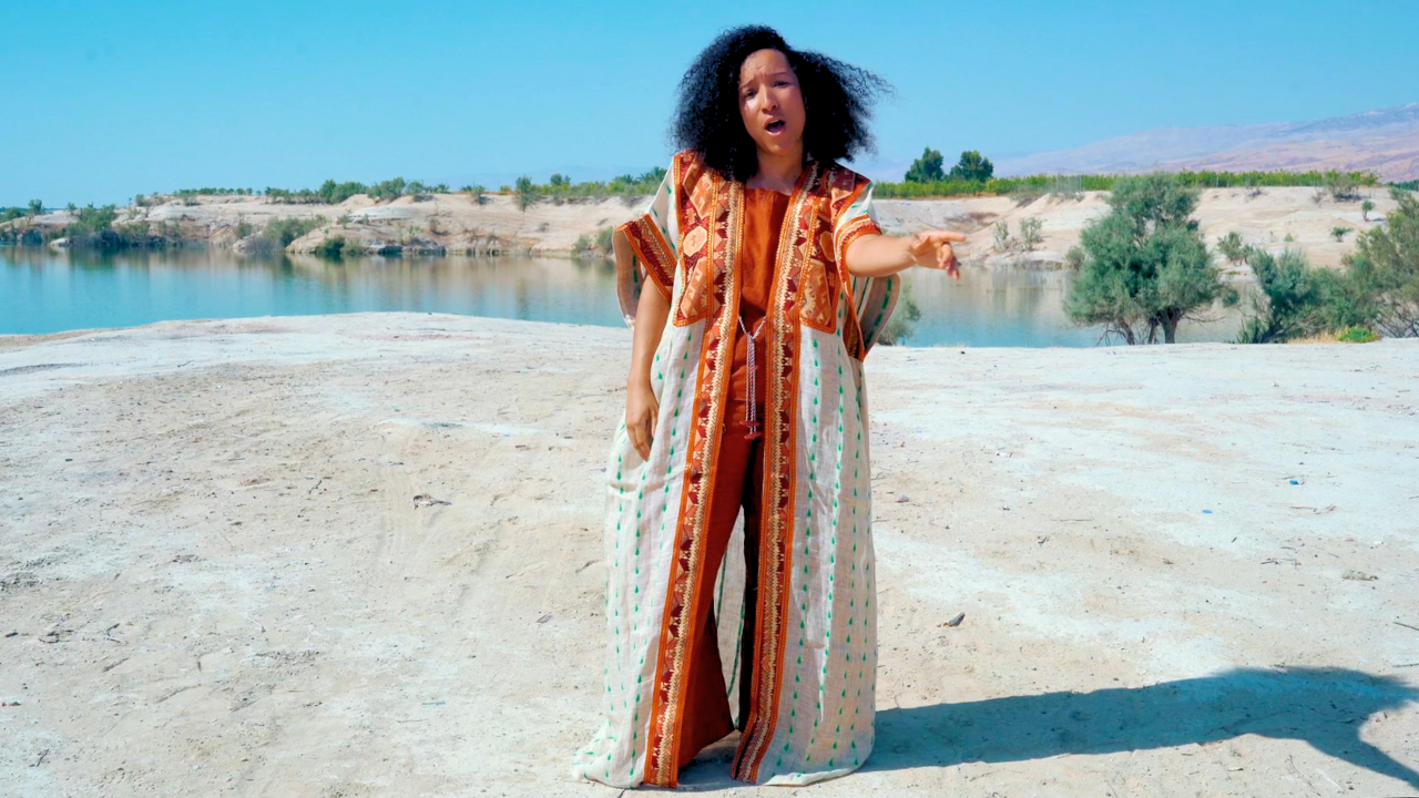 Woman with curly black hair wearing patterned dress standing outdoors near a body of water, with her mouth open and one hand pointing outward.
