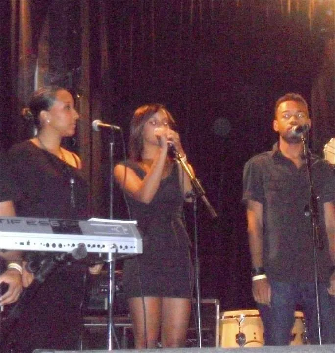 Three young people performing on stage with microphones, a keyboard, and drums, dressed in black, in a dimly lit concert venue.