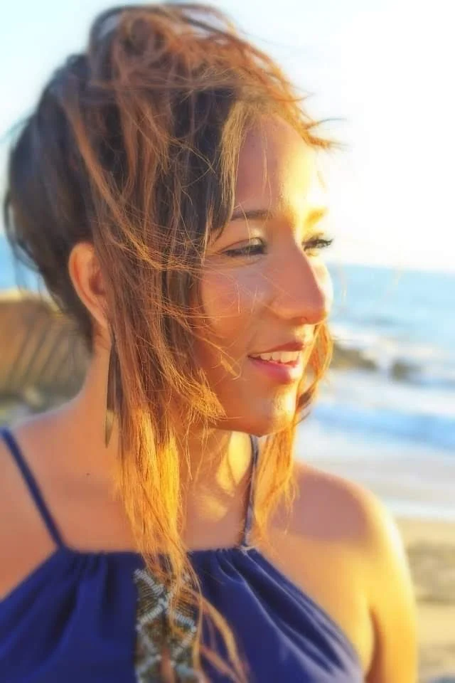 A woman with short, tousled hair smiling on the beach at sunset, wearing a blue spaghetti strap top.