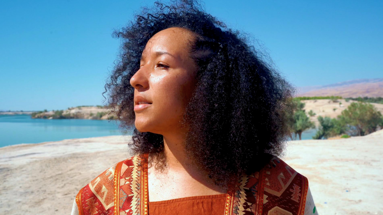A woman with curly black hair standing on a sandy beach near a body of water, with hills and trees in the background under a clear blue sky.