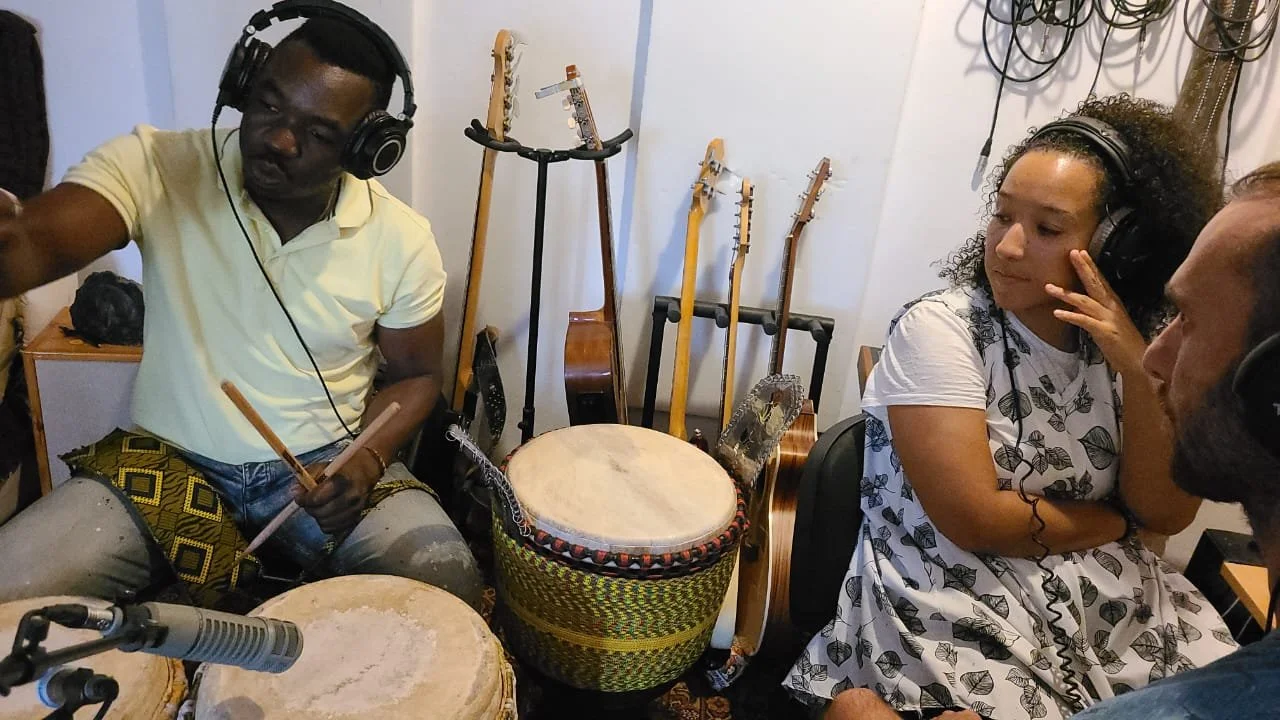 Three people wearing headphones are in a music studio, with drums and guitars in the background. One person is playing drums while the others listen and watch.