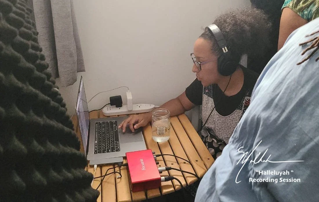 A woman wearing headphones and glasses is sitting at a small wooden table, working on a laptop. There are cables connected to a red audio interface or mixer on the table. A mason jar with water and a charger are also on the table. She is in a room wi