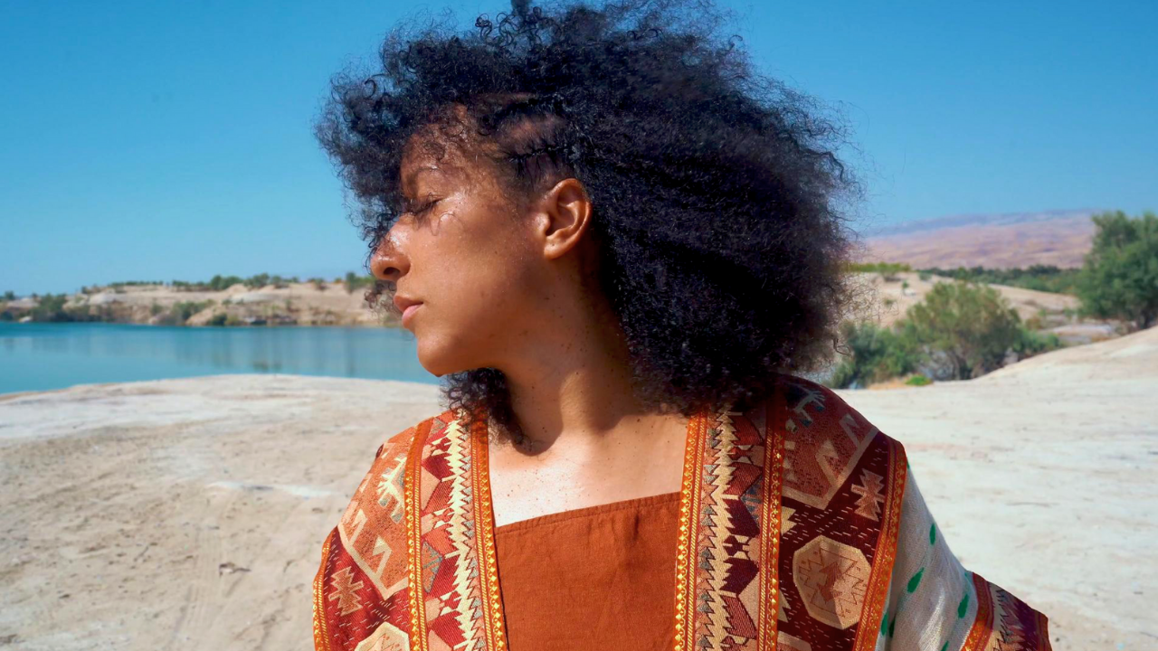 A woman with curly hair and brown skin, wearing a patterned orange and white garment, stands outdoors near a body of water with a scenic landscape of trees, hills, and a blue sky in the background.