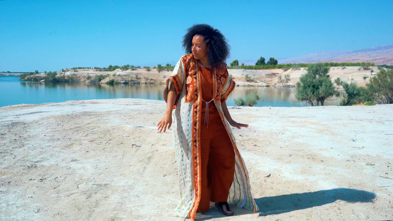 Woman with curly hair wearing a patterned outer garment over orange outfit standing on the sandy bank of a river, with trees and distant hills under a clear blue sky.