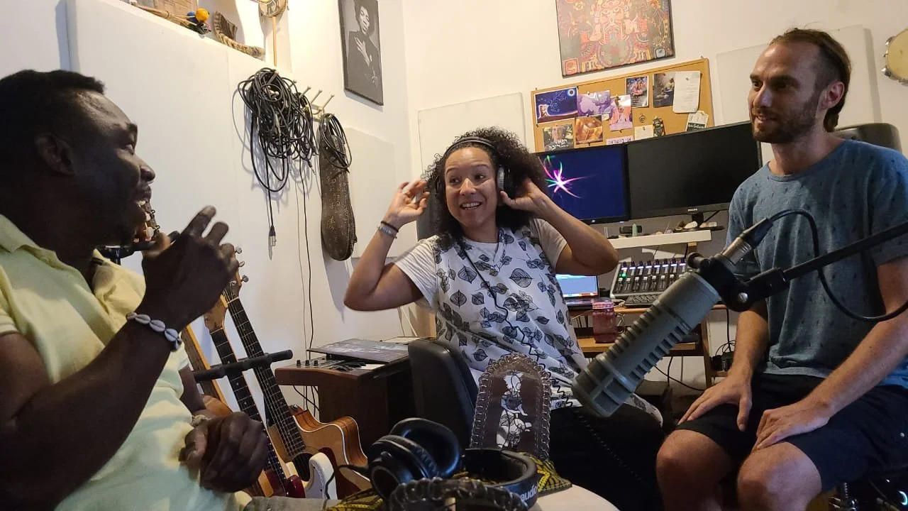 Three people in a recording studio, two men and one woman, engaging in conversation with music equipment around them.