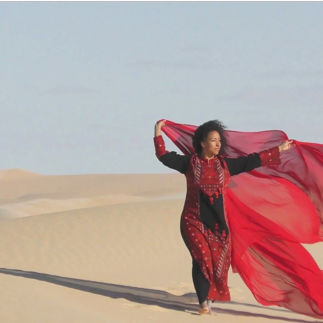 Woman in traditional red and black dress holding a sheer red scarf in a desert landscape with sand dunes and a clear sky.