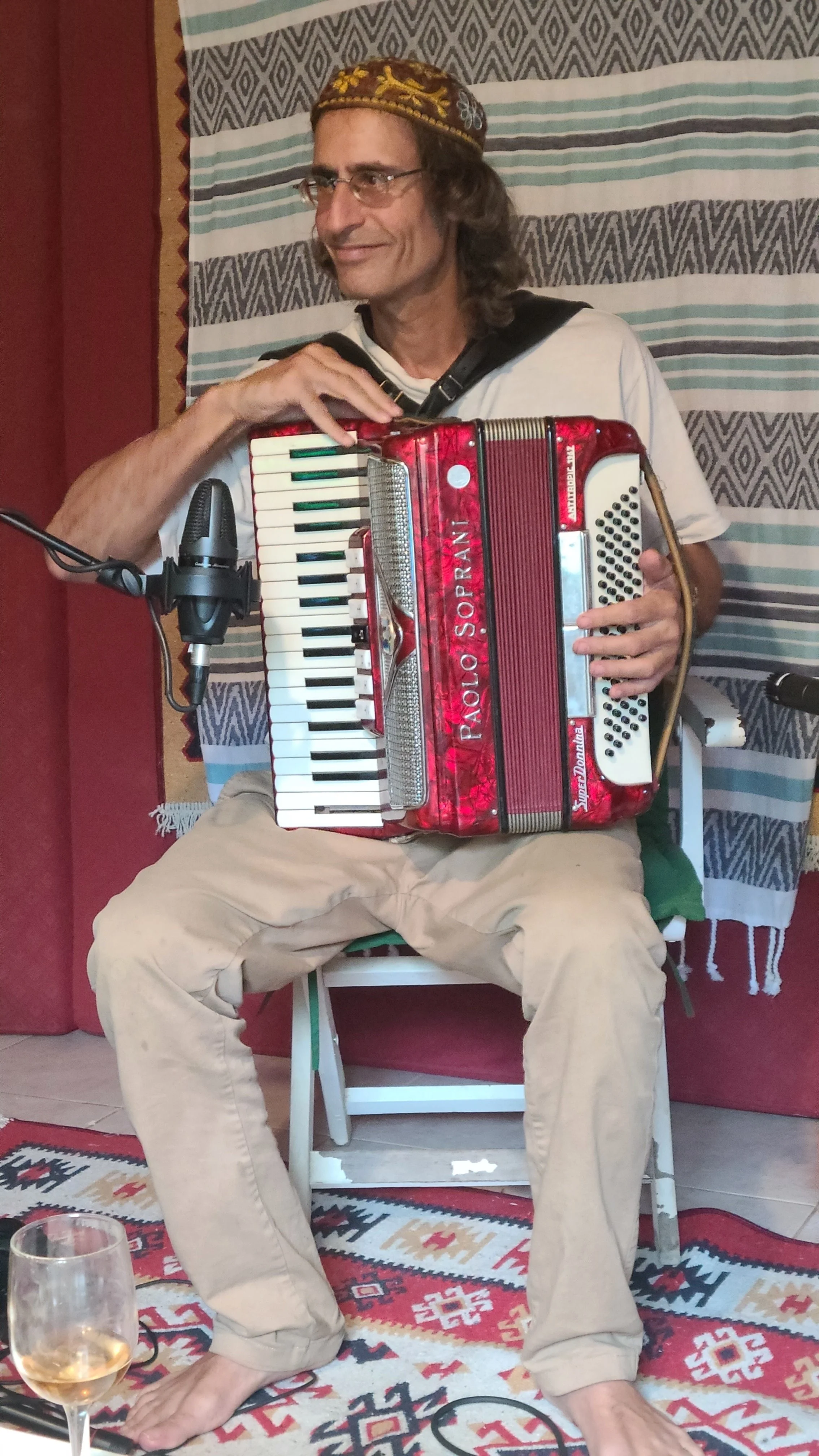A man with glasses, a patterned hat, and long hair playing an accordion while sitting on a white chair. He is dressed in a white t-shirt and beige pants, and in front of him is a glass of white wine on a table. The background features a patterned wal