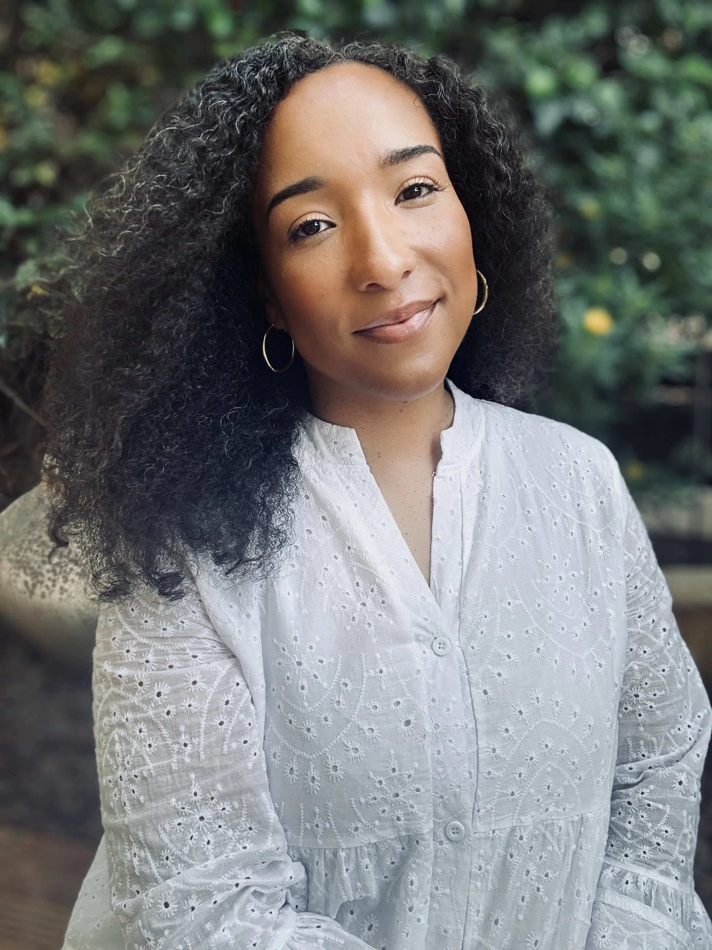 Close-up of a young woman with curly black hair, wearing hoop earrings and a white embroidered blouse, smiling softly in an outdoor setting with greenery in the background.