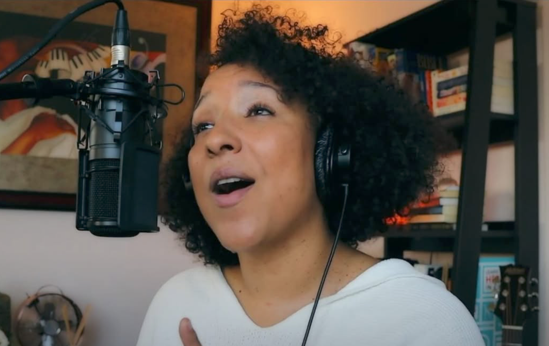 Woman singing into a microphone in a home recording studio with books and artwork in the background.