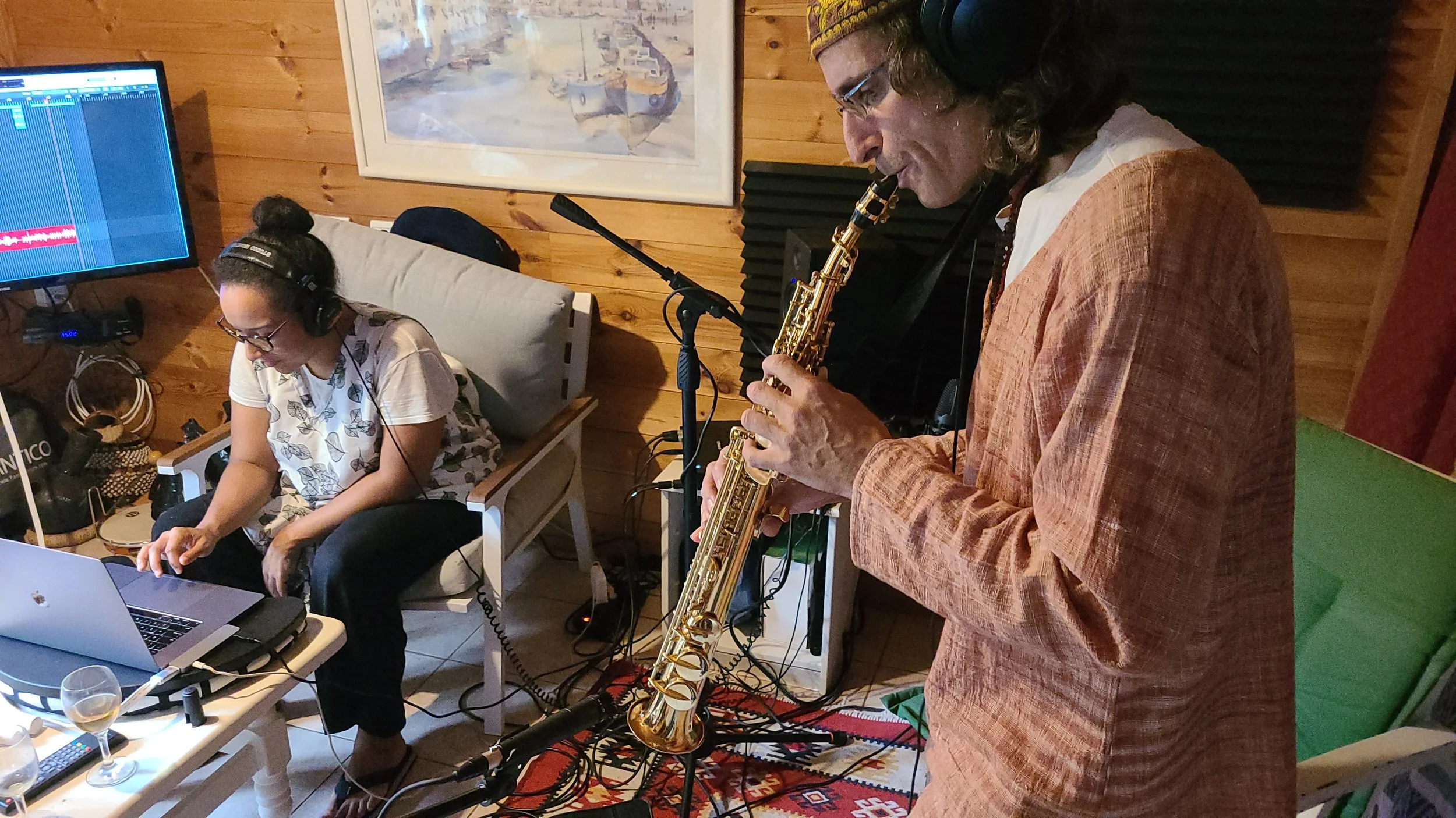 A man playing the saxophone and a woman working on a laptop in a cozy wooden room, with a person in the background wearing headphones and working on electronic equipment.