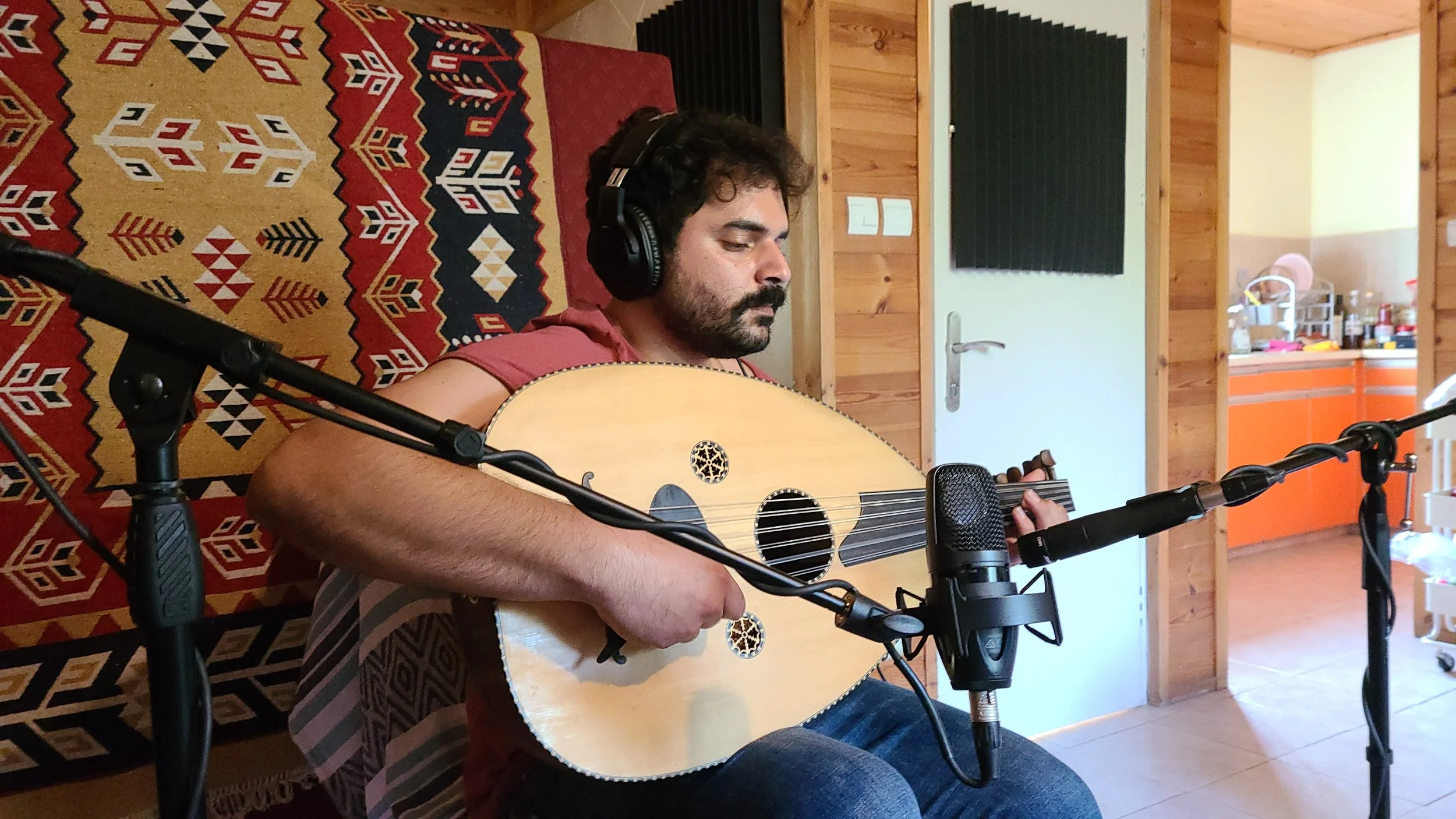 A man with dark curly hair and a beard wearing headphones playing an acoustic guitar in a cozy wooden room with a patterned rug and soundproofing panels.